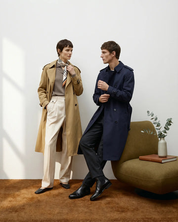 A fashion editorial featuring classic Burberry trench coats. The female model wears the iconic honey-colored trench with a matching check scarf, while the male model poses in a tailored navy blue trench coat. High-end minimalist aesthetic.