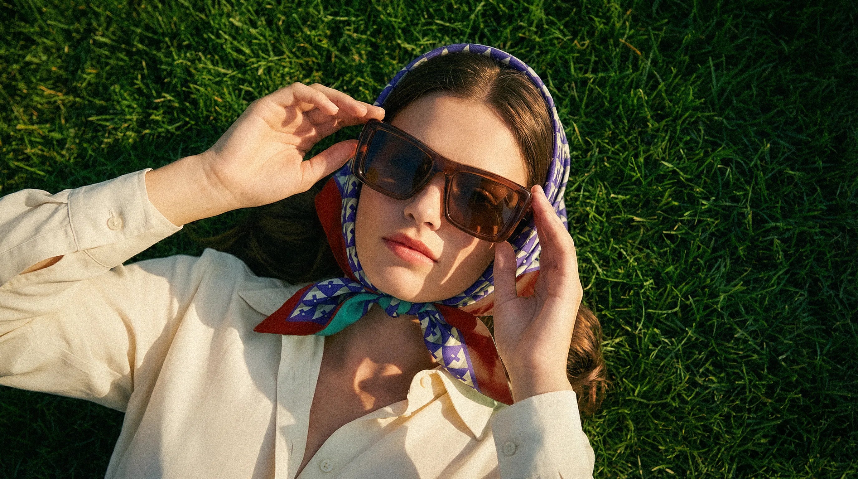 Woman wearing Saint Lauren sunglasses and a colorful Miu Miu silk foulard with light cream shirt lying on grassslider_item_GDBQHh