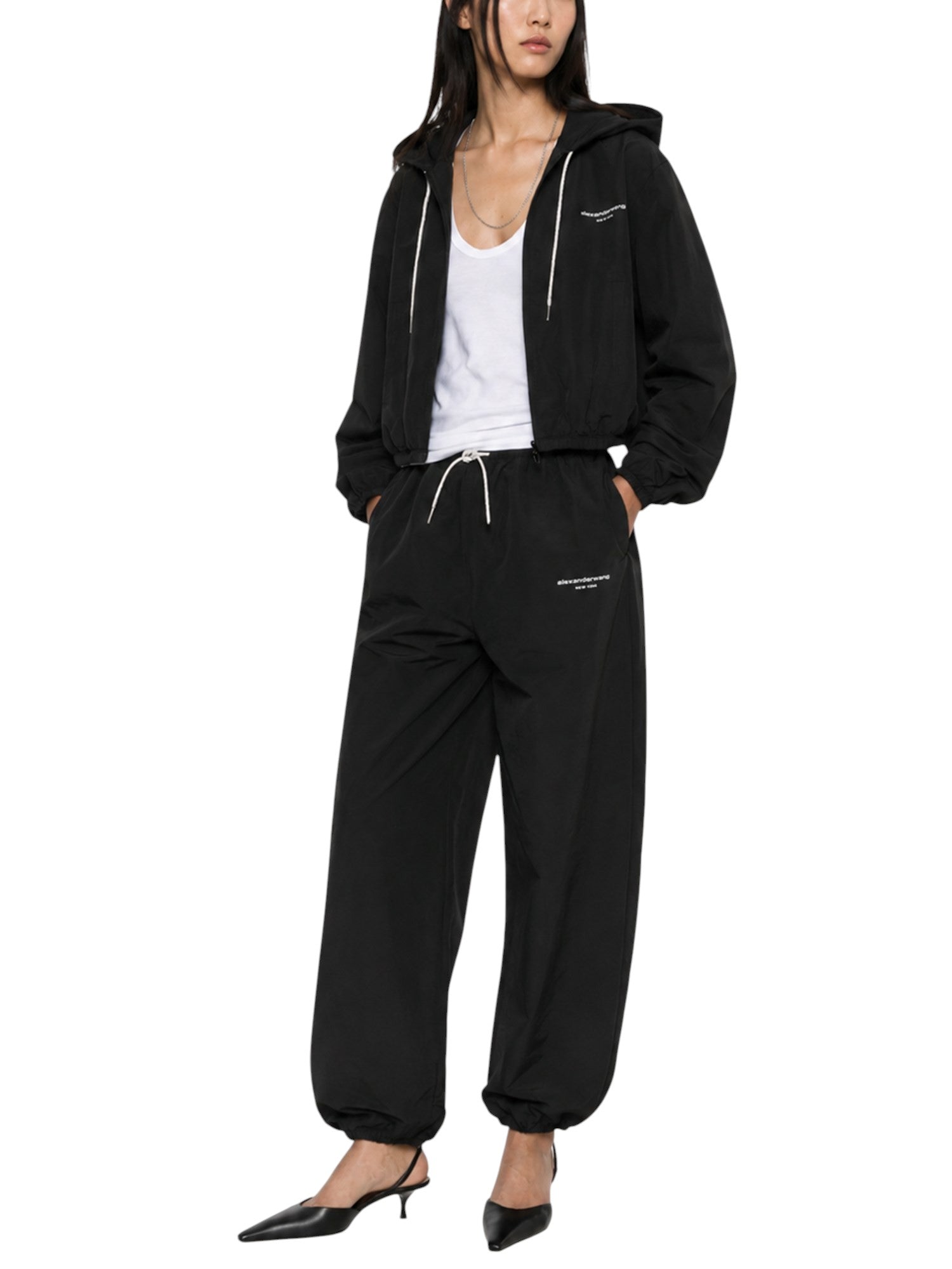 Model wearing Alexander Wang black technical track pants with a matching cropped hooded jacket and white tank top.