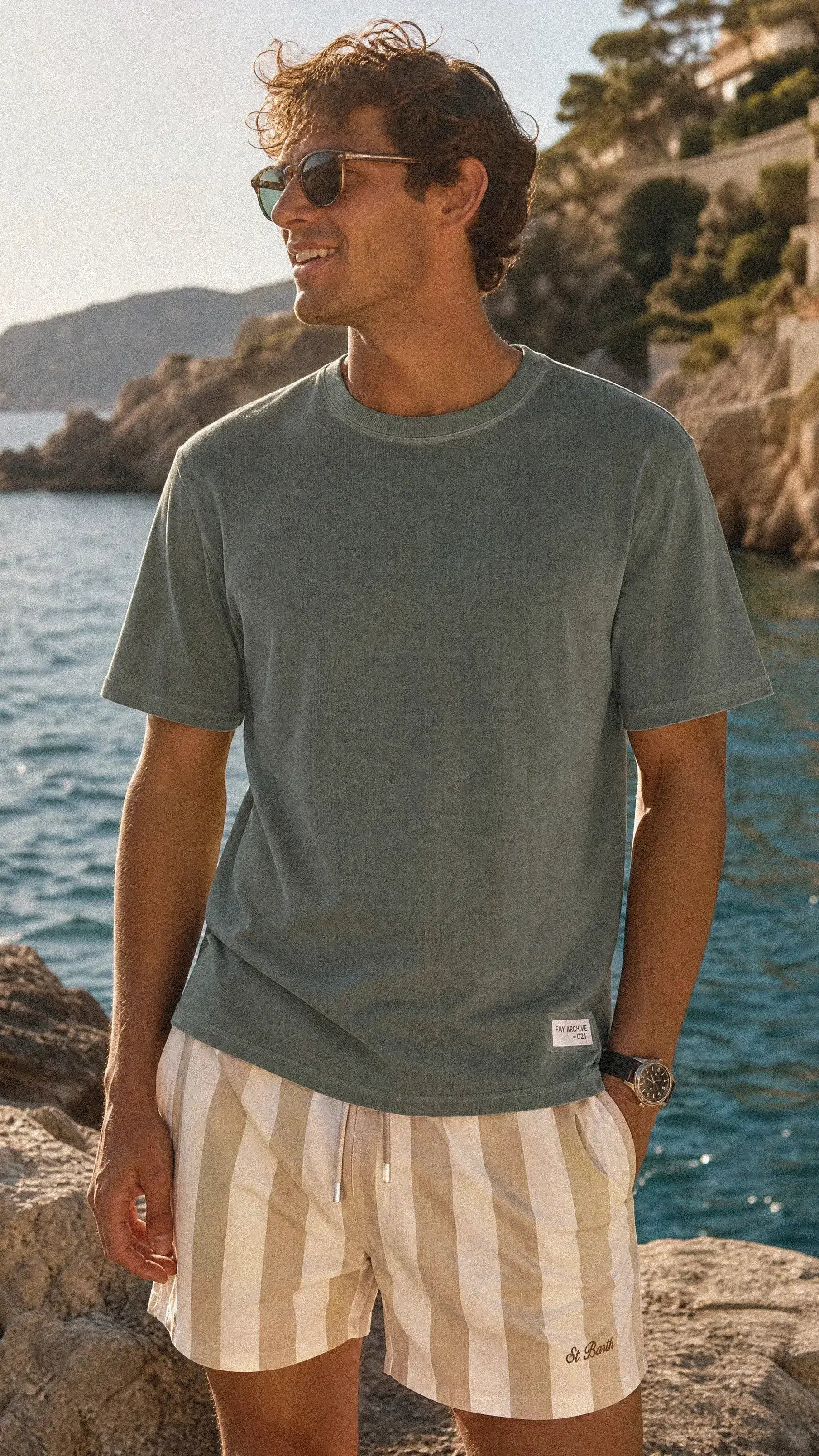 A man wearing a muted sage FAY green crewneck t-shirt and cream and MC2 SAINT BARTH beige vertically striped swim trunks, standing on a rocky coastline with the sea and buildings in the background.