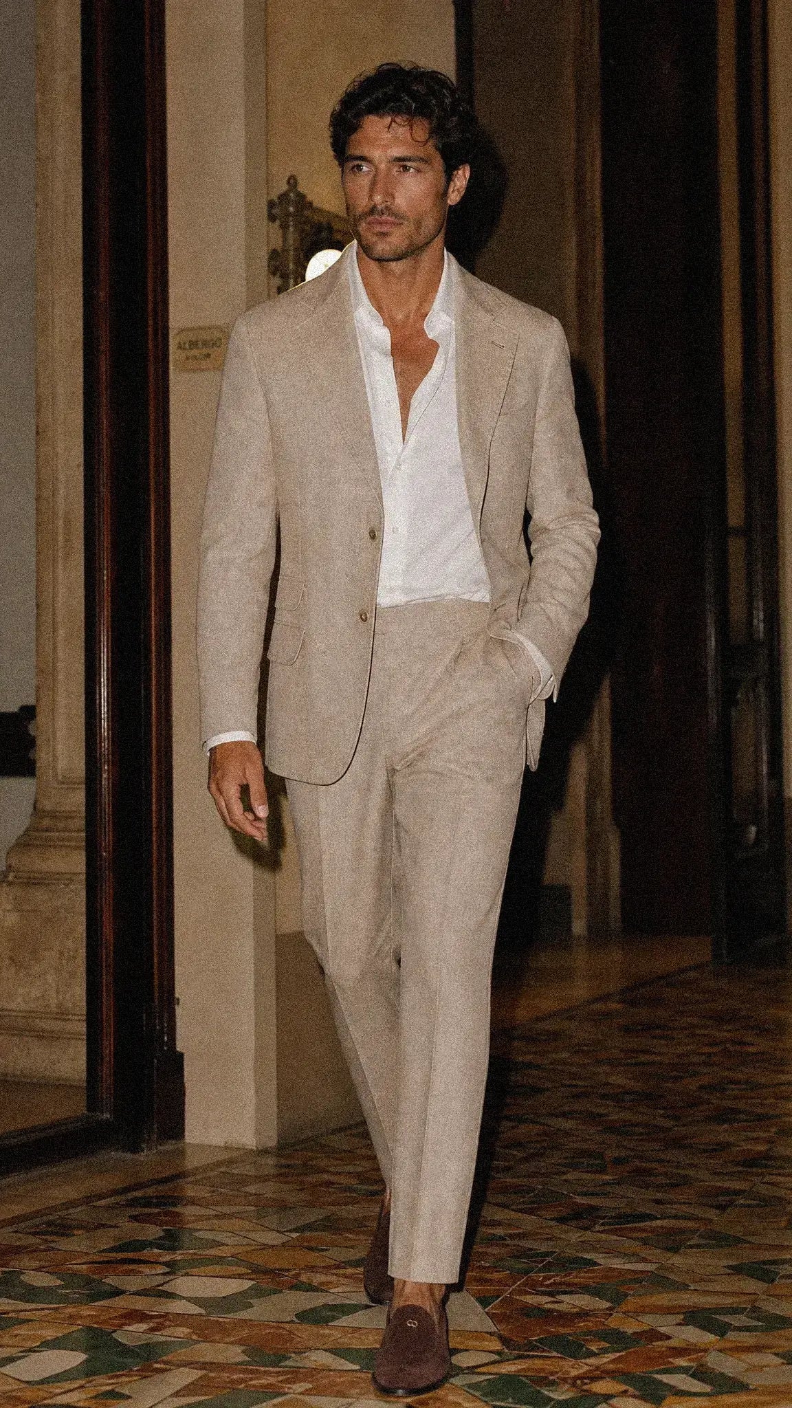 A man in Ceremony Scene walks down a hallway wearing a Brunello Cucinelli light beige linen suit with a white open-collar shirt and  Dior brown loafers.