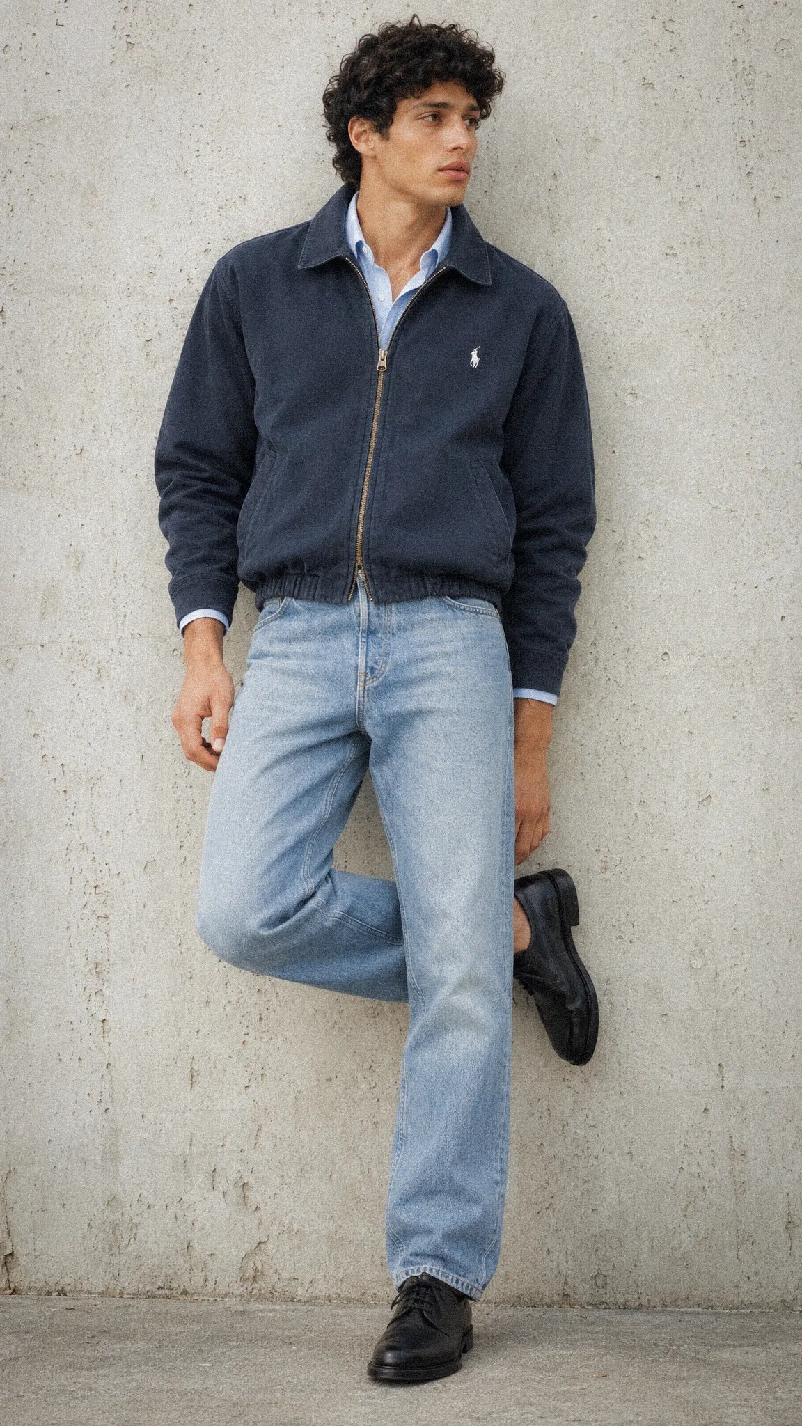 A man wearing a Polo Ralph Laurent navy blue zip-up jacket with a small embroidered polo player logo on the chest, light-wash blue jeans, a light blue collared shirt, and black shoes, leaning against a textured concrete wall.