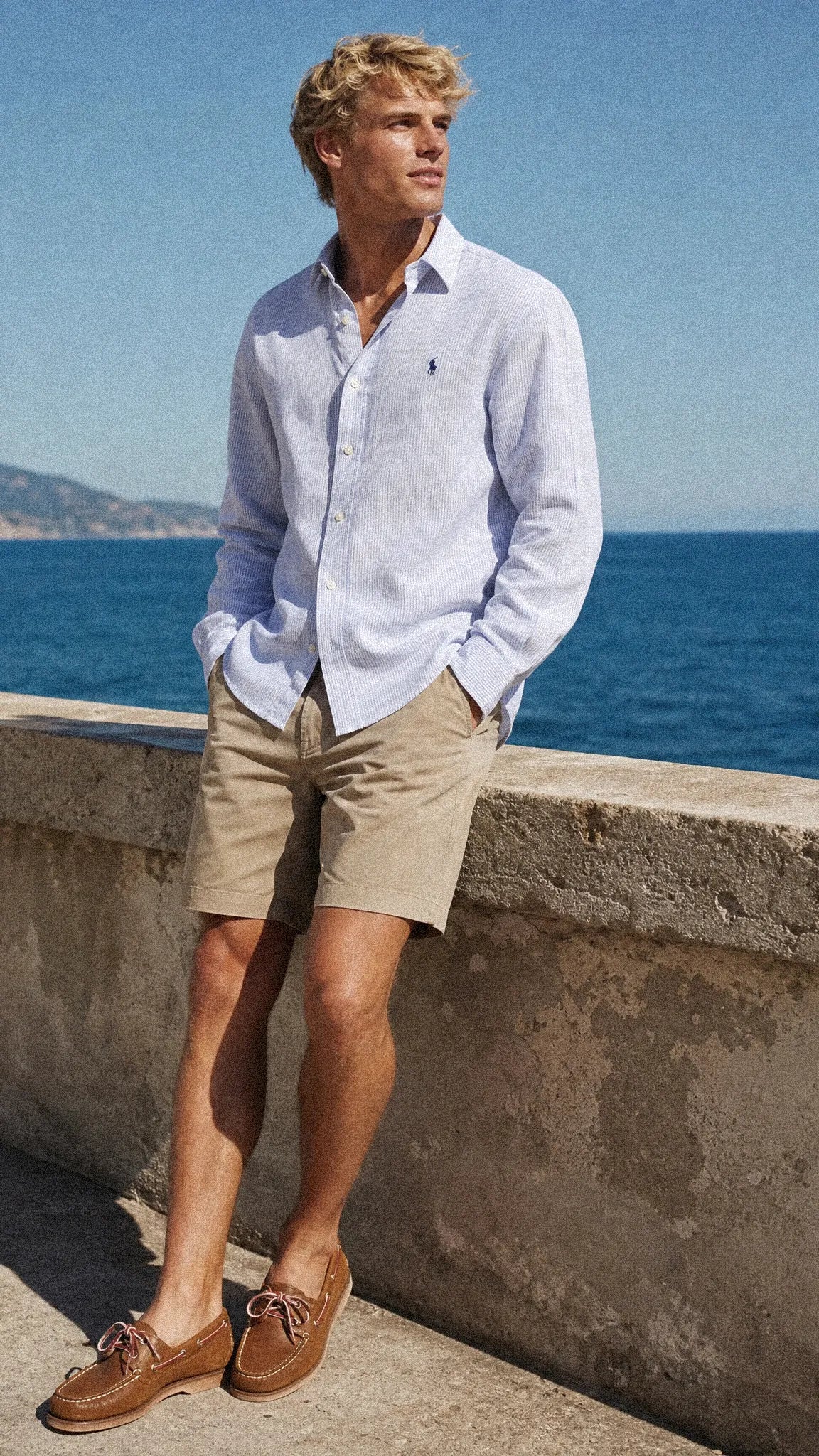 A man stands outdoors by the sea wearing a Ralph Lauren  light blue long-sleeve button-down shirt, khaki shorts, and brown boat shoes.